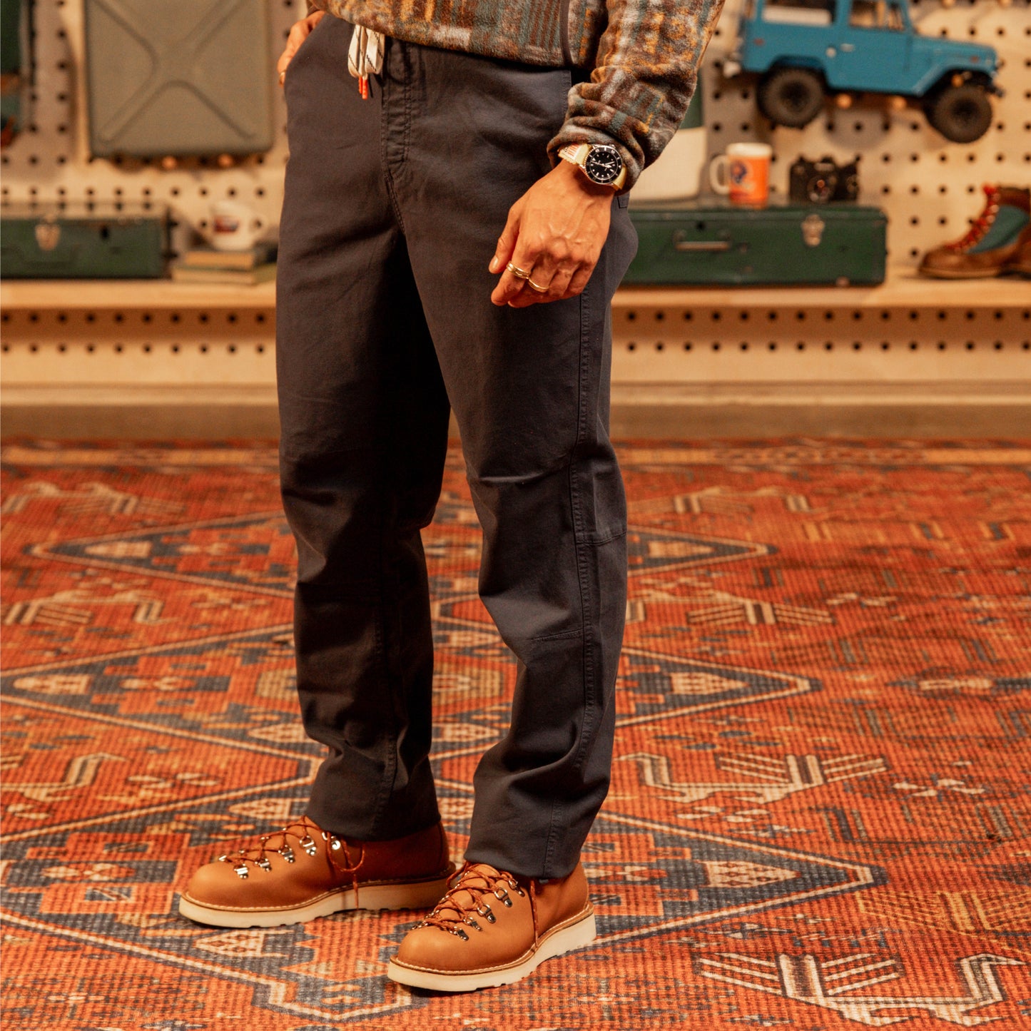 Person wearing dark pants and brown boots standing on a patterned rug with a workshop background.